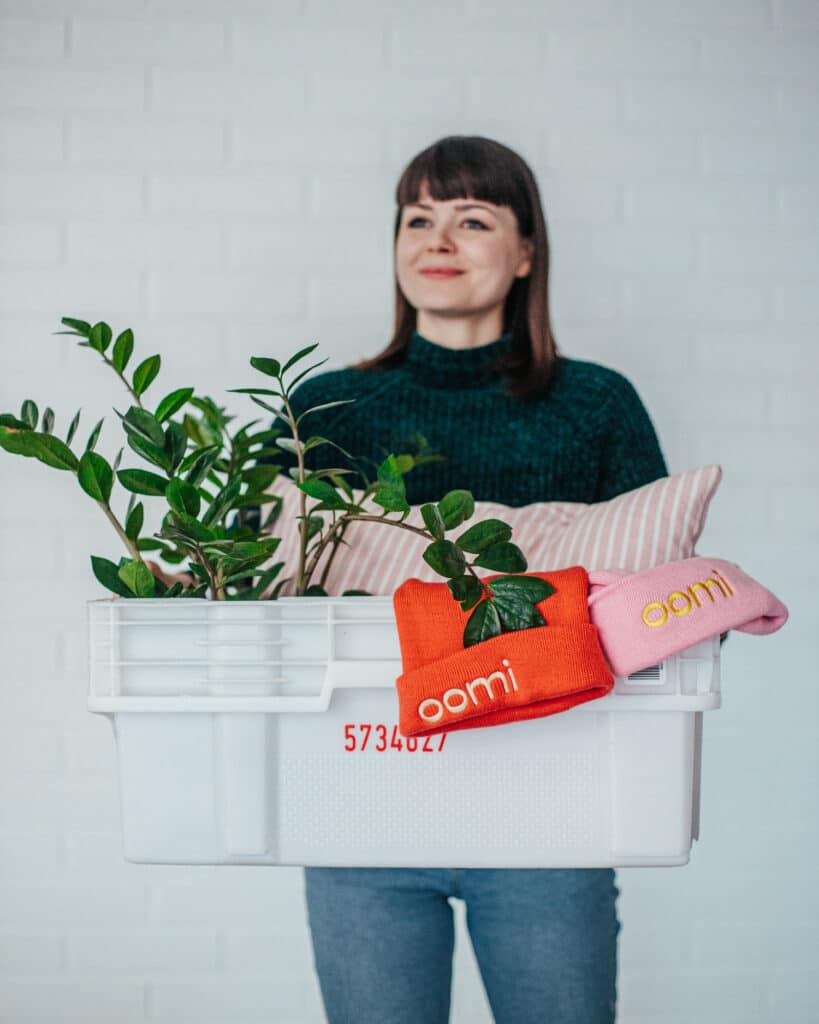 Nainen kantaa muuttolaatikkoa, jonka sisällä on huonekasvi, tyyny ja kaksi pipoa.